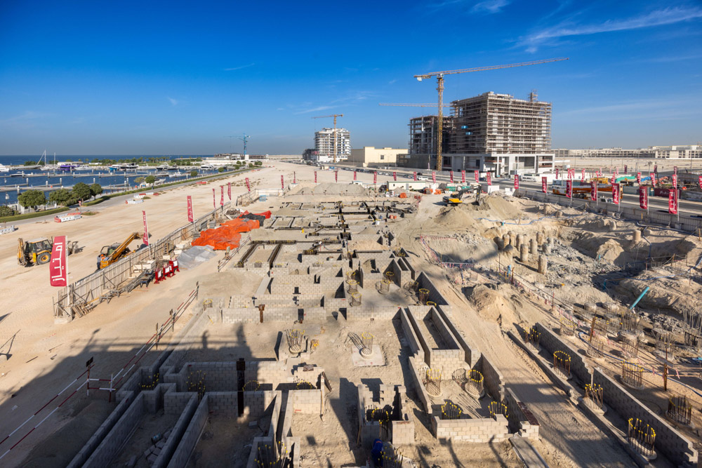 Cotier House 2 By Imtiaz Construction site with heavy machinery, building foundations, and cranes under a clear blue sky near a waterfront.